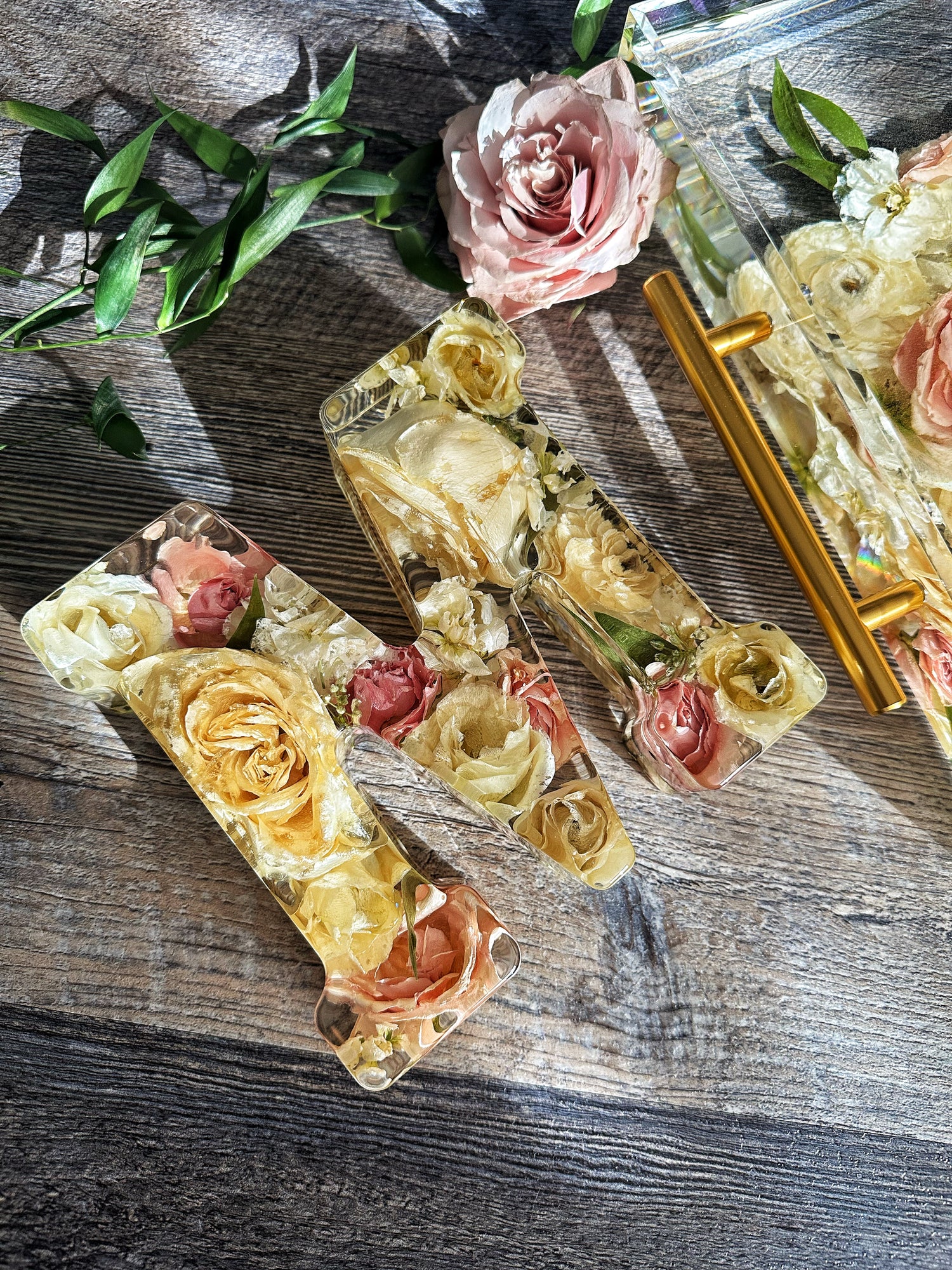 Decorative resin letter with pressed flowers on a wooden surface with a rose and green leaves.