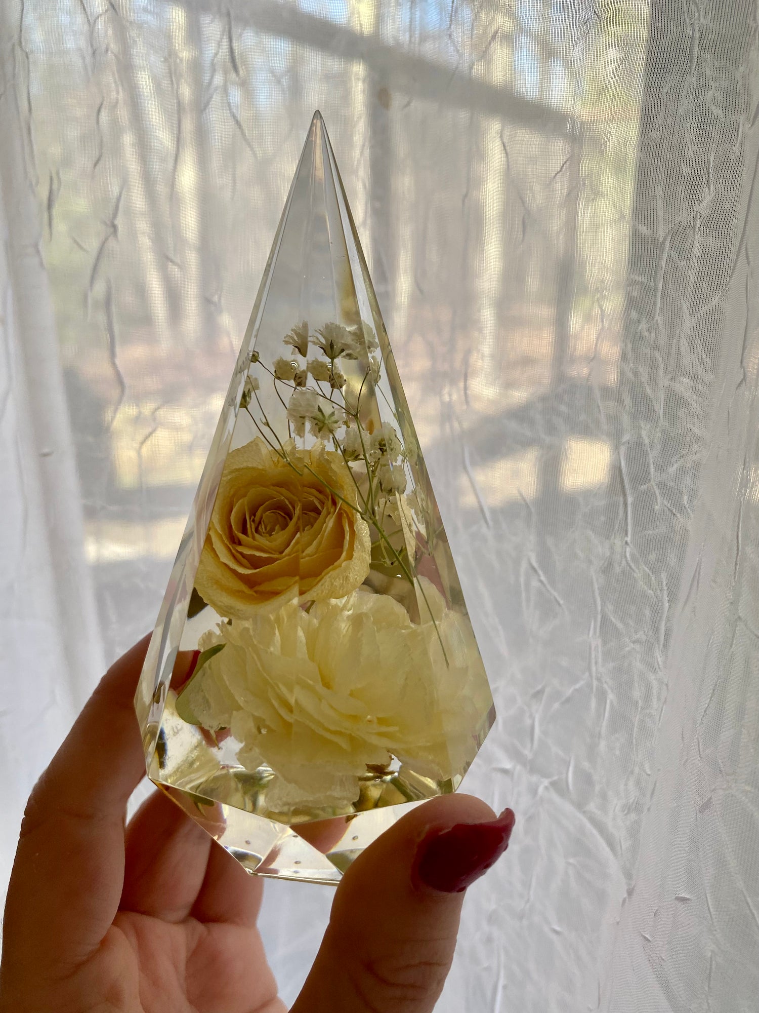 Hand holding a clear teardrop-shaped container with preserved flowers against a window backdrop.