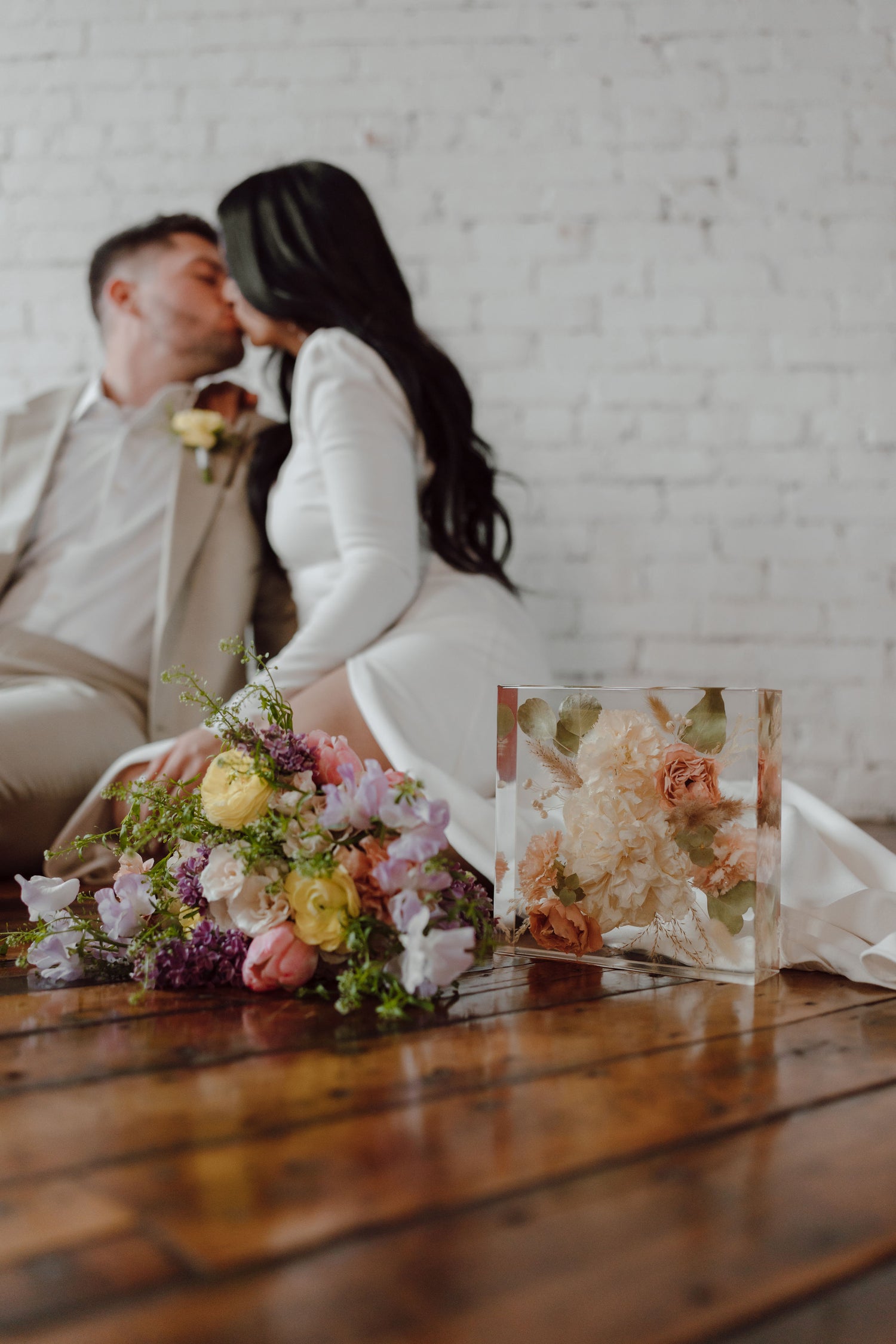 bride and groom sitting together with a preserved bouquet of flowers on a wooden floor.